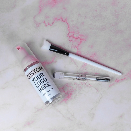 An image showing a custom 3-piece lash aftercare kit with logo, including 3 bottles with foam cleanser, lash wands, and cleansing brushes, displayed on a marble surface with pink veining.
